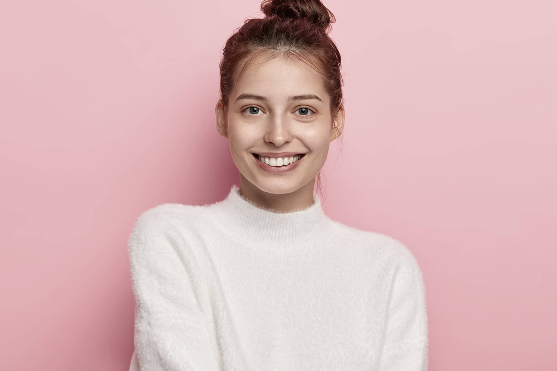 tender feminine woman with blue eyes smiles pleasantly has toothy smile wears white comfortable sweater looks directly camera isolated pink background